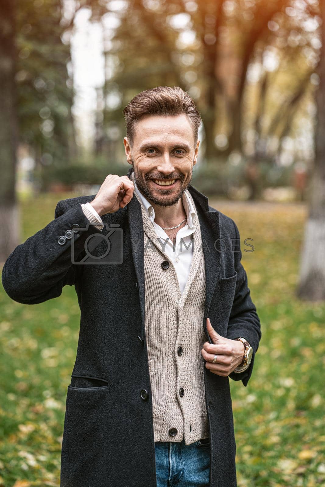 Royalty free image of Handsome smiling happy man straightens the sides of his dark blue coat. Young freelancer man stands on the street in an autumn coat and cardigan under it looking happy at camera by LipikStockMedia