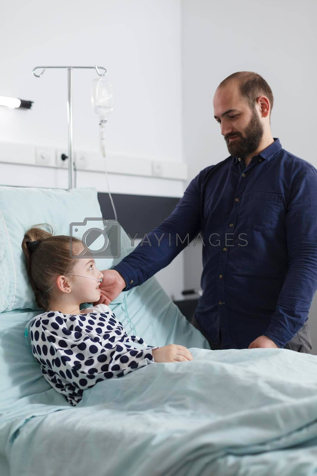 Careful supportive father caress sick little daughter while laying in hospital patient bed. Thoughtful lovely parent offering ill child patient emotional support while resting in patient room.