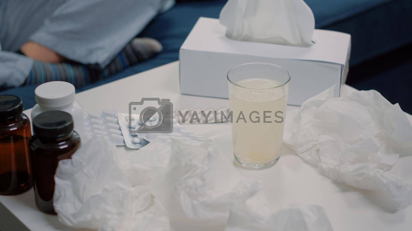 Close up of effervescent vitamin drink in glass of water on table with tablets of medicaments, bottles of pills, and tissues to cure cold and flu symptoms. Person with healing beverage