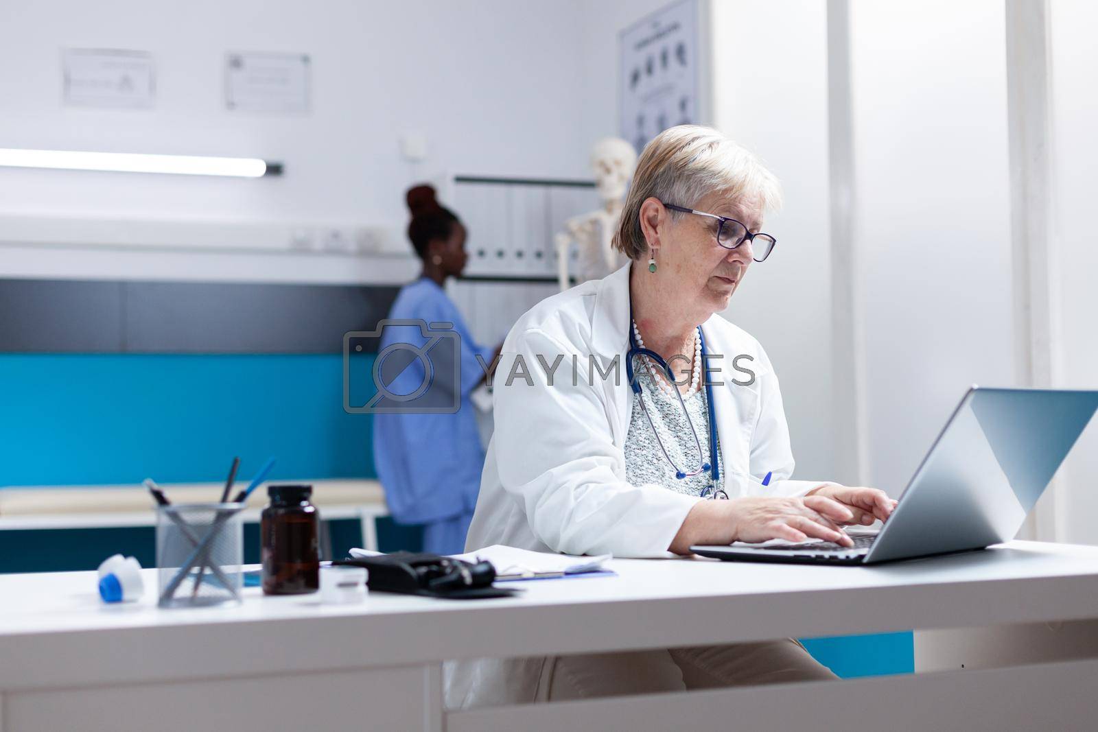 Senior doctor using laptop in cabinet to search for healthcare treatment. General practitioner looking at computer screen to work on prescription medicine against disease diagnosis.