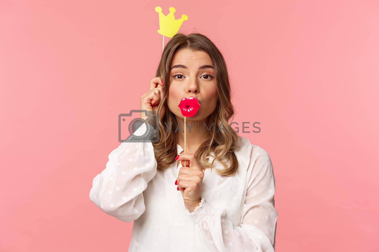 Spring, happiness and celebration concept. Close-up portrait of funny and cute, silly blond girl in white dress, holding big lips mask and crown, partying and having fun, pink background.