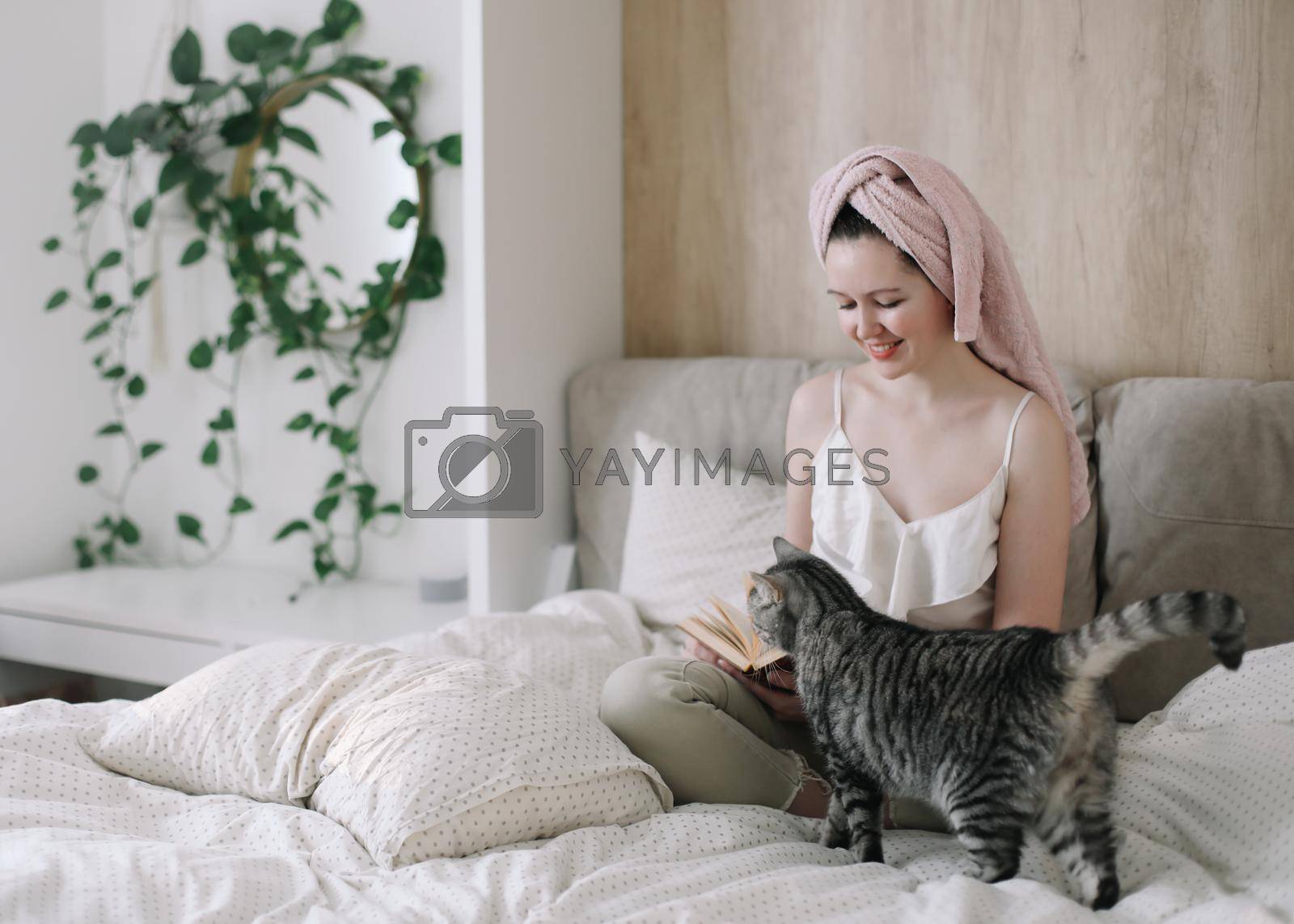 Young woman and her adorable cat reading book and resting in bed. Lazy weekend at home with loved pet concept. Close up, copy space, interior background. Love, togetherness and pets indoors.