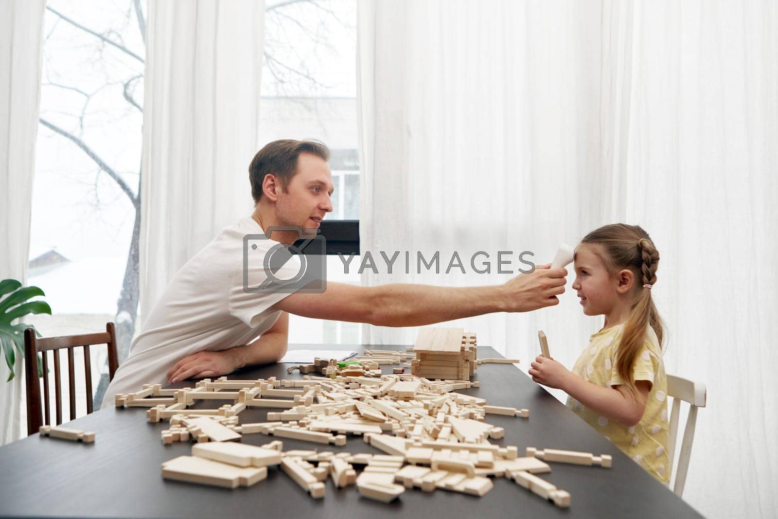 Side view of dad checking body temperature with infrared forehead thermometer of daughter sitting at table and playing with wooden blocks