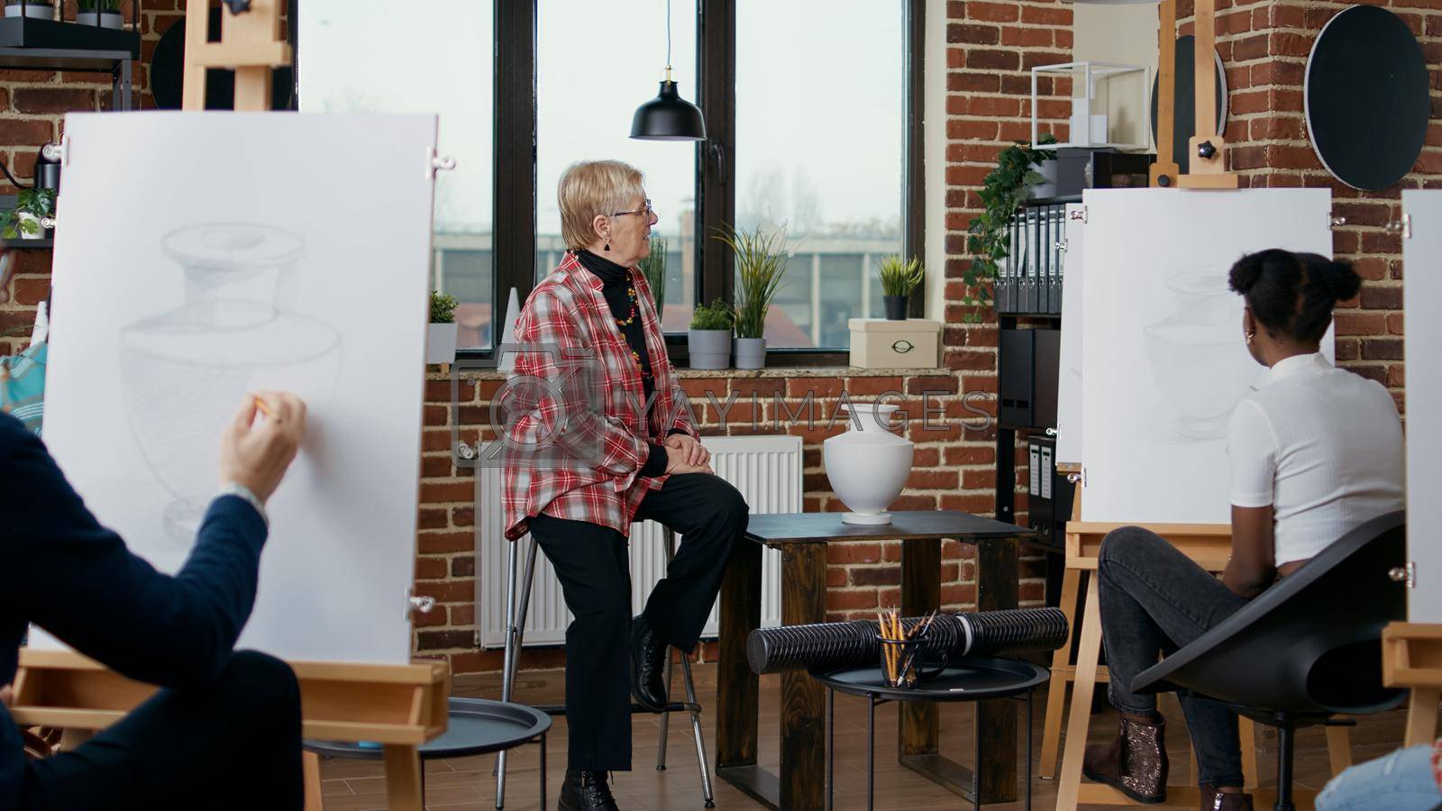 Senior woman explaining artistic vision to people in art class, teaching multiethnic group of students to draw masterpiece on canvas. Teacher using vase model to show drawing inspiration.