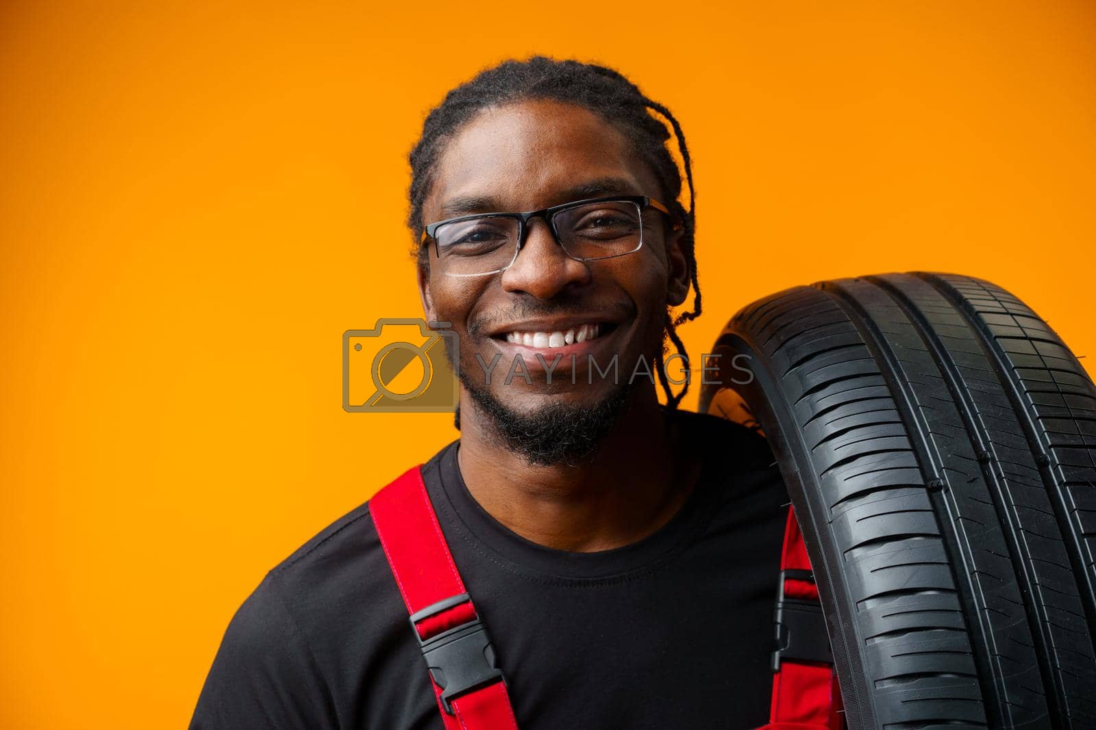 Royalty free image of African american car service worker with car tyre against yellow background by Fabrikasimf