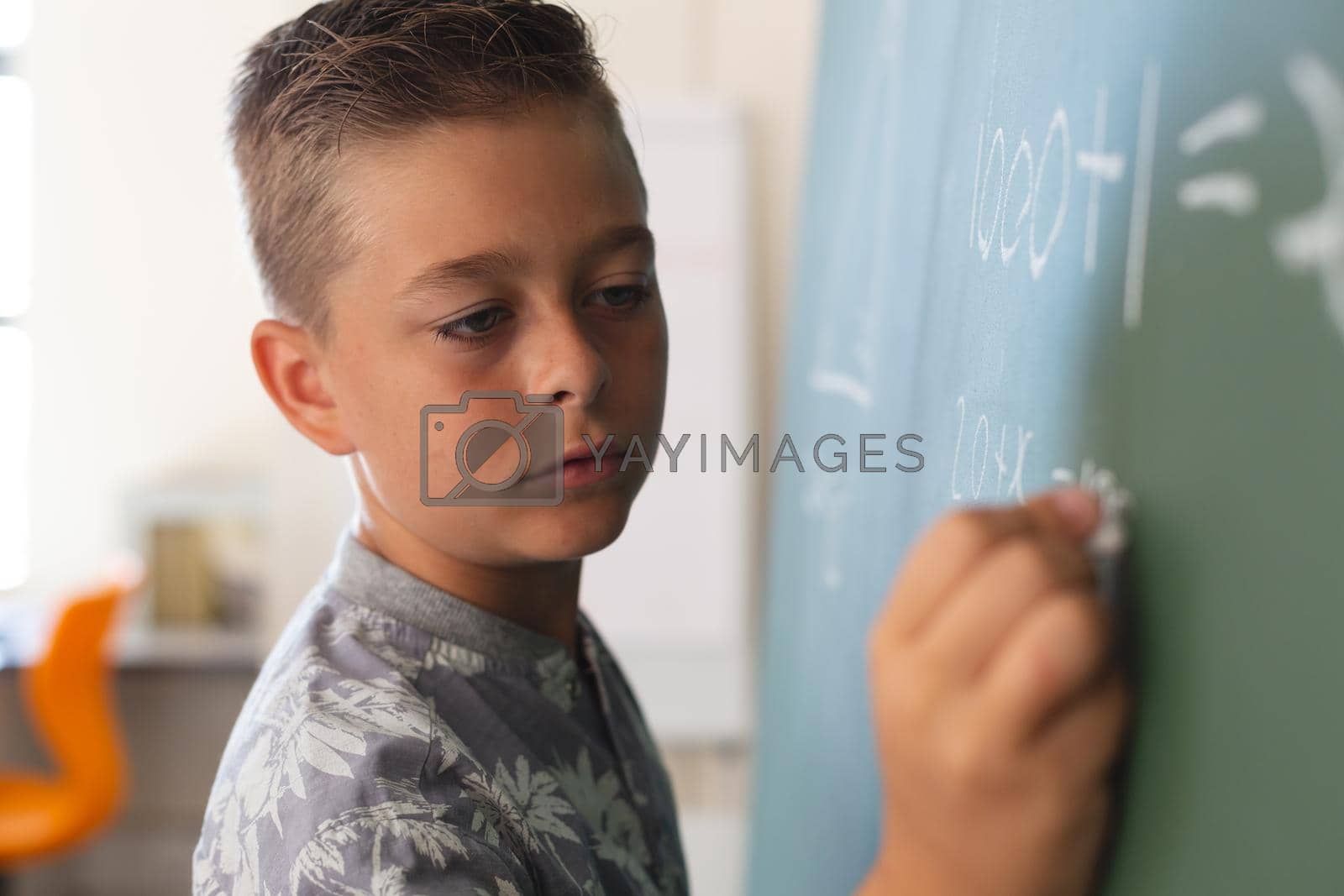Royalty free image of Caucasian boy standing at chalkboard writing in classroom during maths lesson by Wavebreakmedia