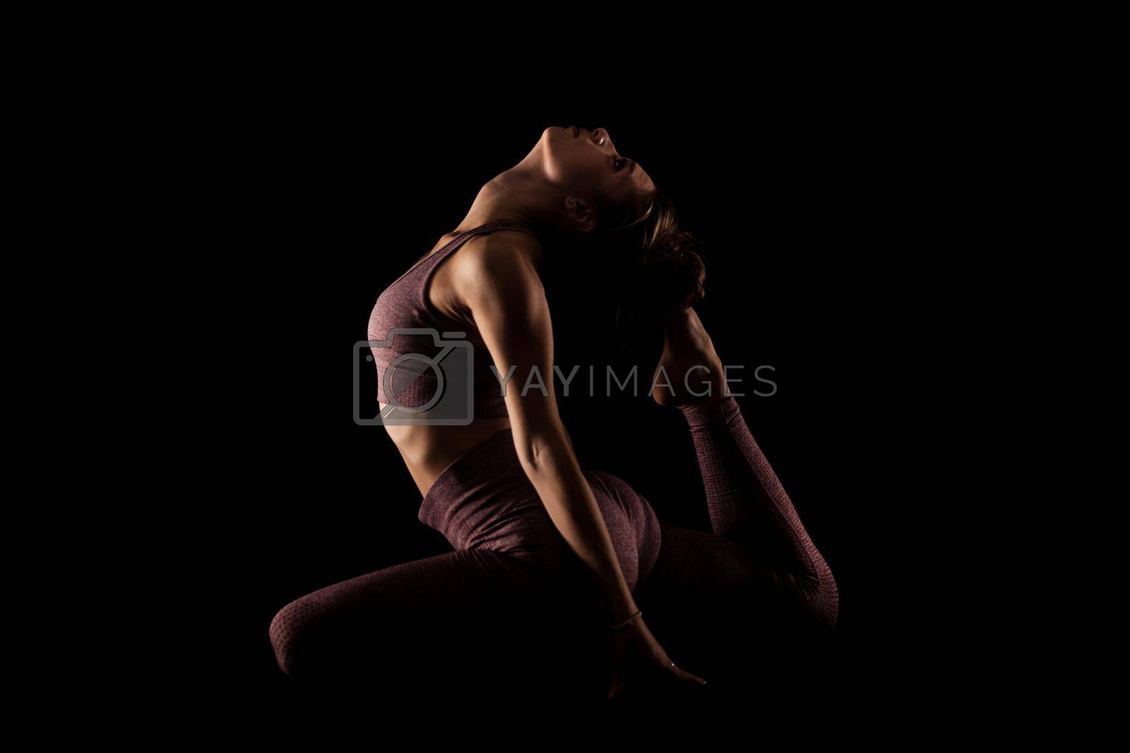 Fit woman practicing yoga poses. Side lit half silhouette girl doing exercise in studio against black background.