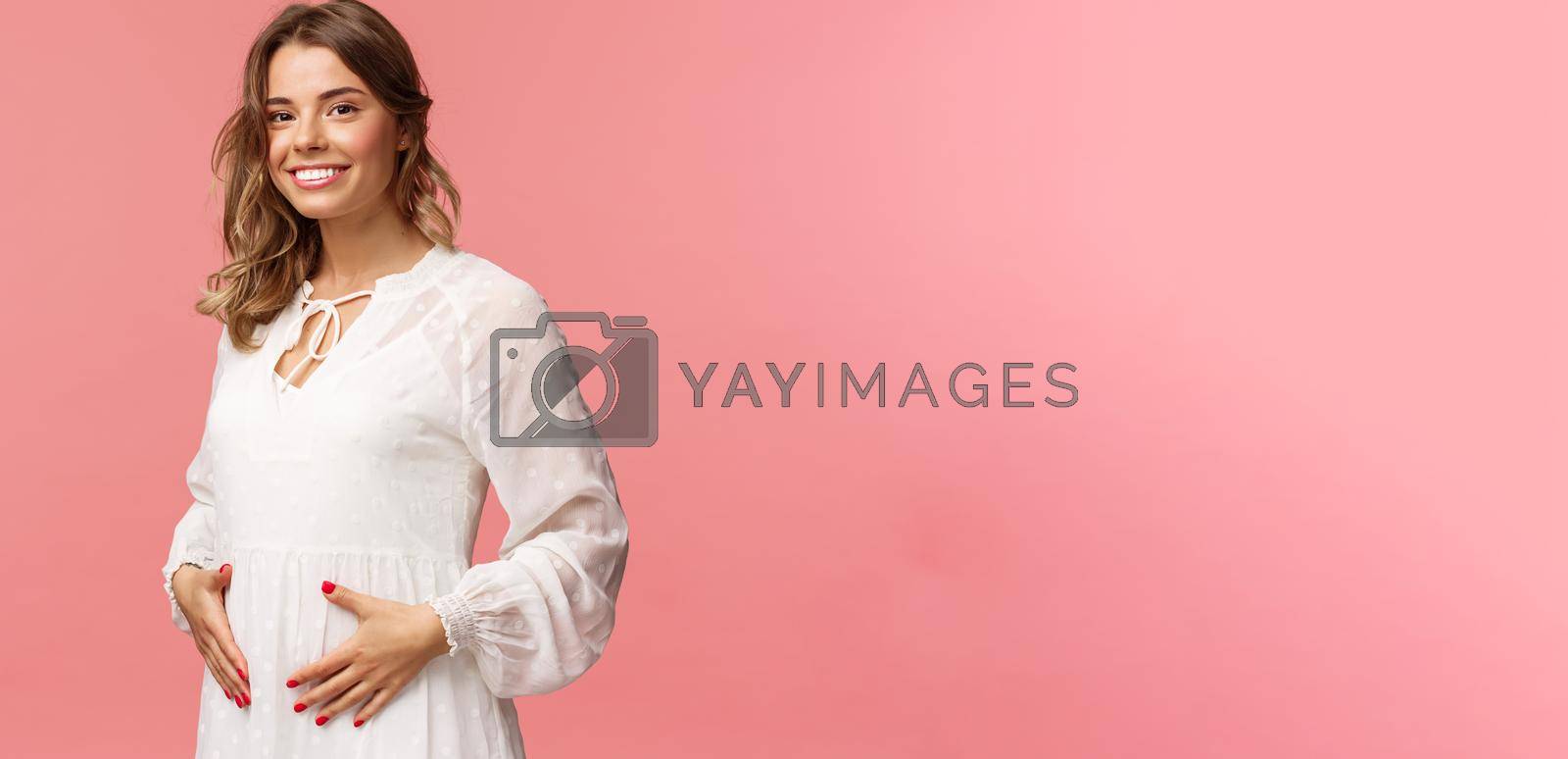 Maternity, women and beauty concept. Tender, cute smiling blond woman in white dress feeling happiness and love, touching belly as expecting child, being pregnant, pink background.