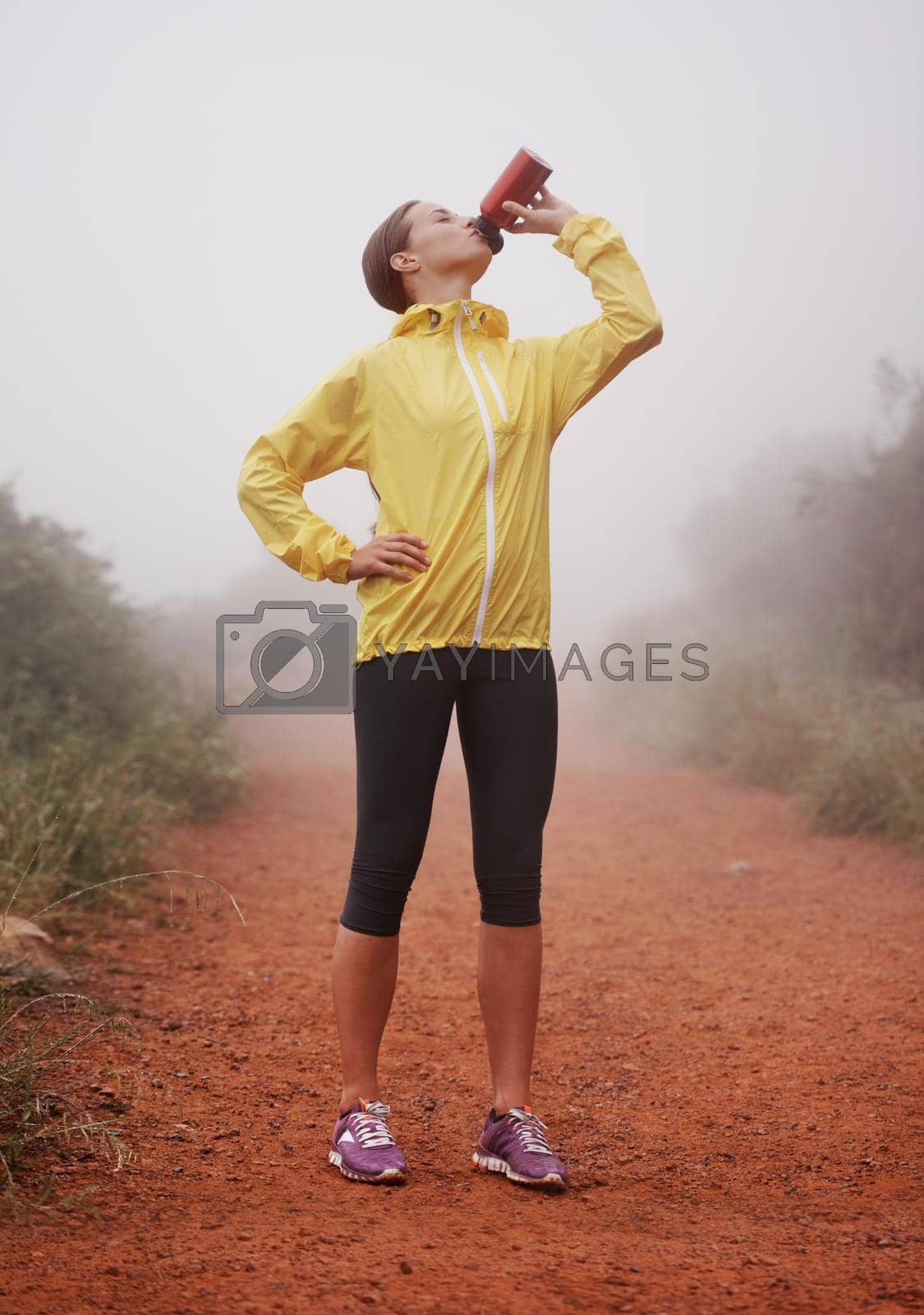 A young female runner drinking from her water bottle while standing in a forest trail.