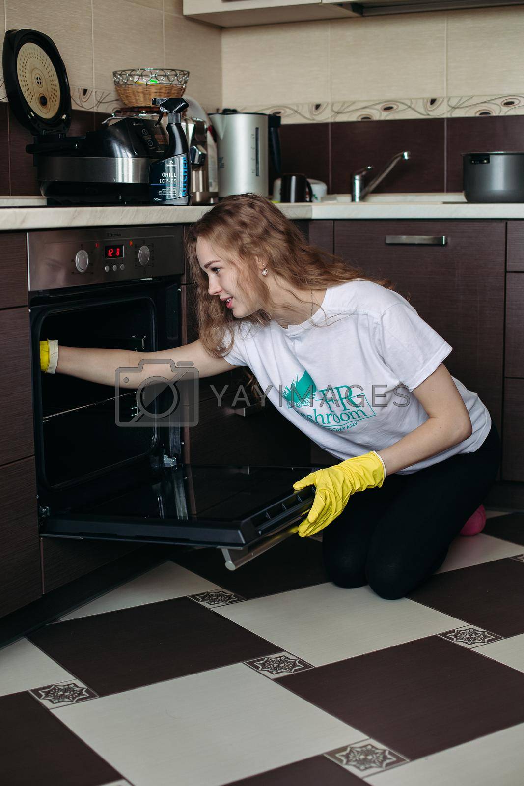 Belarus Minsk 06 29 2018 : Young woman in yellow protective gloves holding head by hands, looking dirty oven at home kitchen. Brunette girl with curly hair washing oven on kitchen holding hand inside.