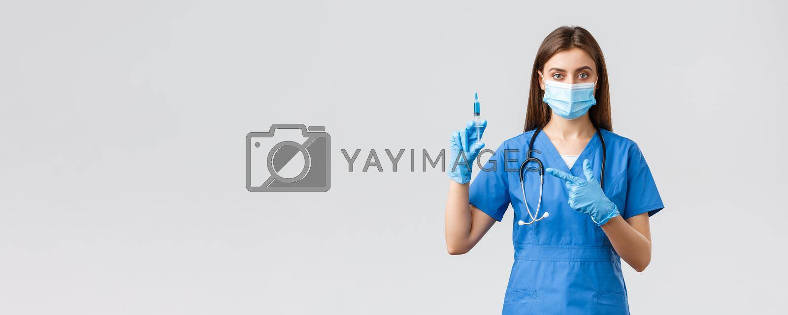 Covid-19, preventing virus, health, healthcare workers and quarantine concept. Serious female nurse or doctor in blue scrubs, medical mask, pointing at syringe with coronavirus vaccine.