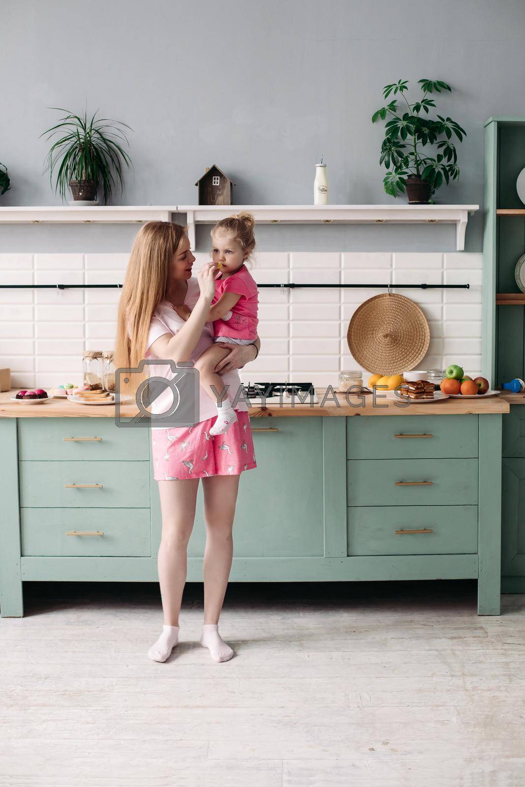 Full length of beautiful smiling woman in pyjamas holding her lovely daughter on hands and feeding her with fruit for breakfast. Modern kitchen.