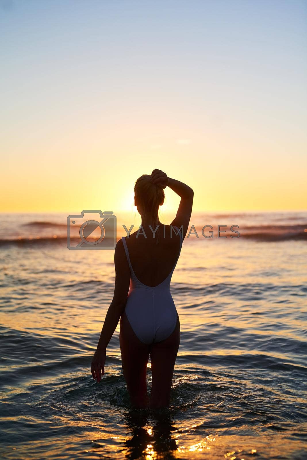 Rearview shot of a young woman standing in the water at sunset.