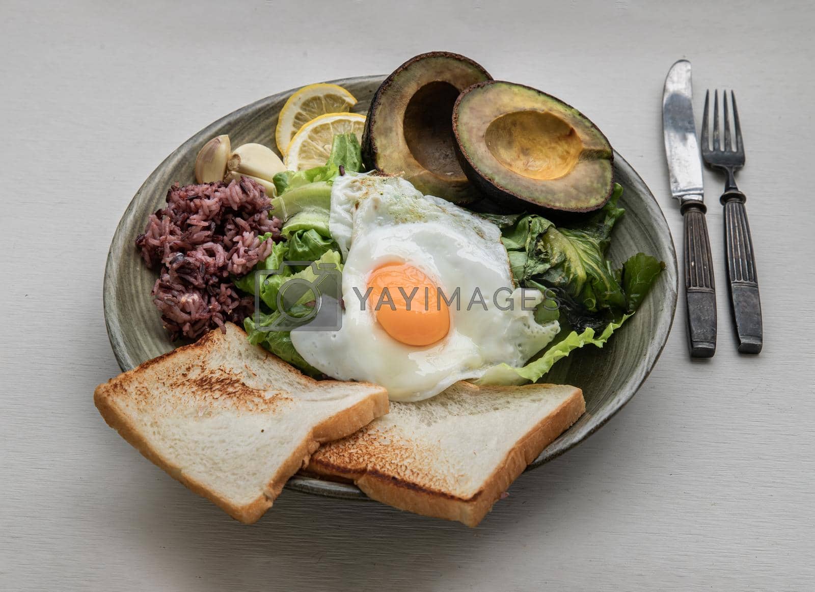 Royalty free image of Breakfast with Fried Egg to put it on top of Fresh vegetables, Rice berry, Breads, and Garlic with Avocado cut in half and lemon sliced on Ceramic plate.  by tosirikul