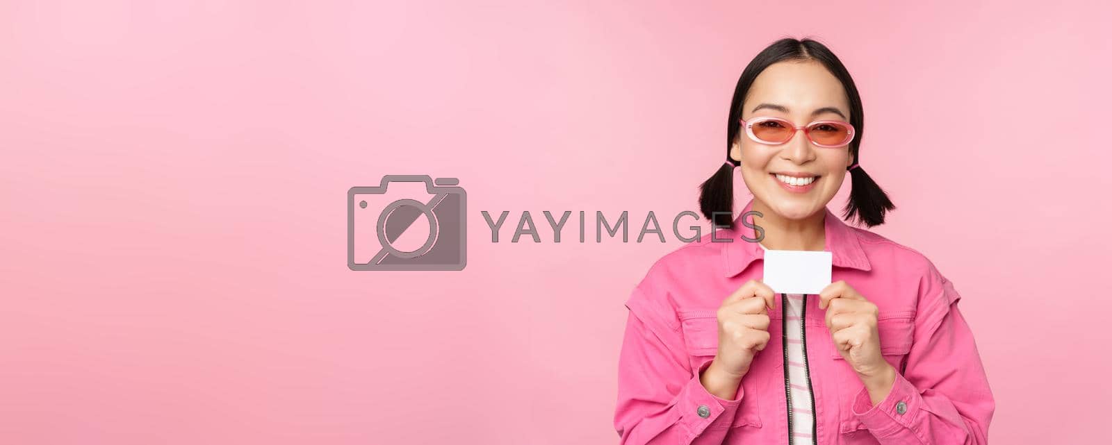 Royalty free image of Portrait of stylish, modern asian girl, shows discount, credit card and look pleased, paying contactless, concept of shopping, standing over pink background by Benzoix