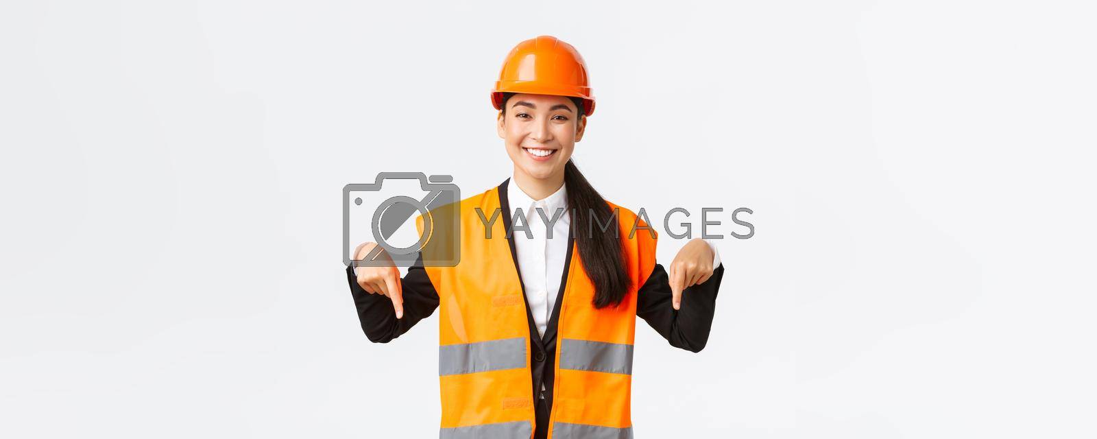 Royalty free image of Smiling cheerful construction manager, asian female engineer in safety helmet and reflective clothing showing announcement. Woman project manager pointing down, standing white background by Benzoix
