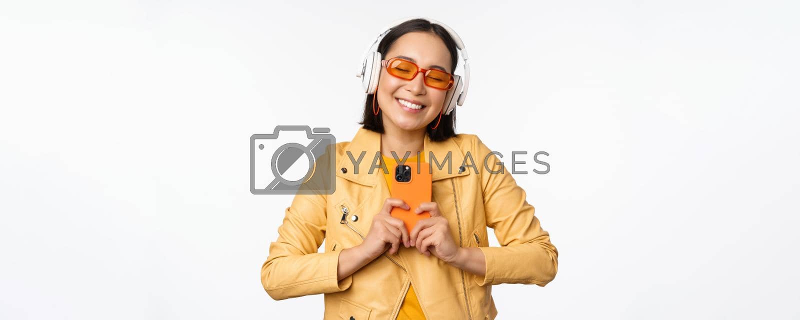 Technology concept. Stylish asian girl in headphones, holding smartphone, dancing and singing, listening music, standing over white background.