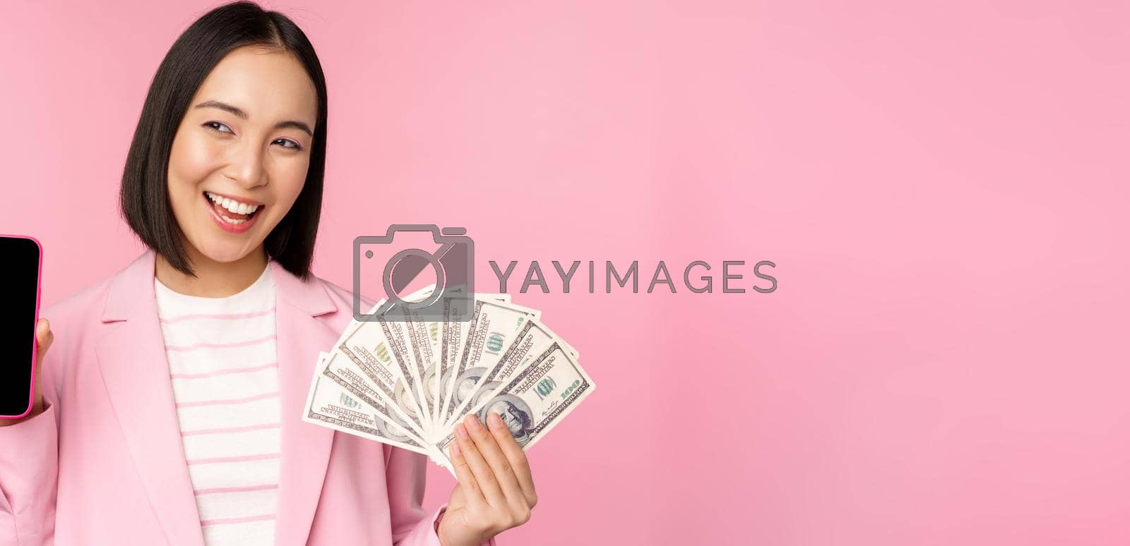 Royalty free image of Successful young asian businesswoman showing money, cash dollars and smartphone screen, smiling pleased, pink background by Benzoix