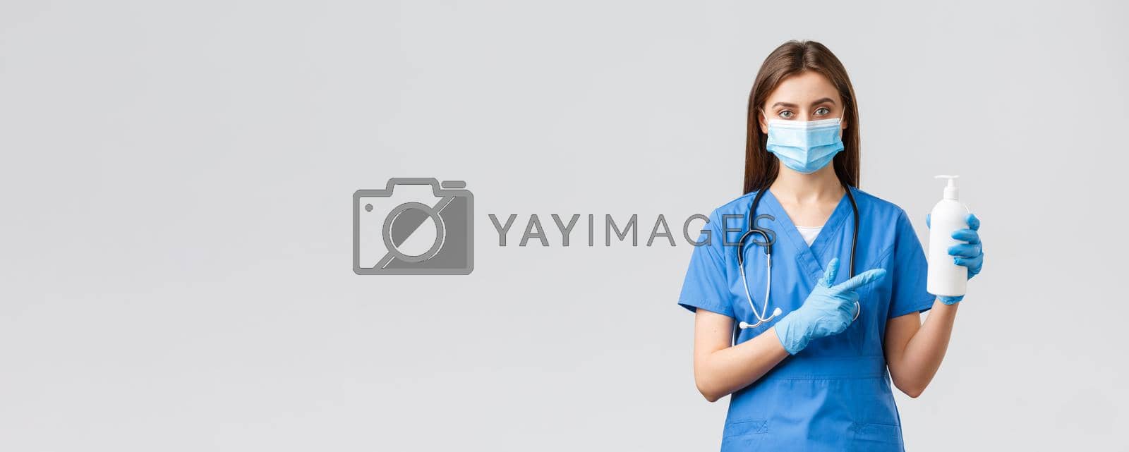 Covid-19, preventing virus, health, healthcare workers and quarantine concept. Young pretty female nurse or doctor in blue scrubs, medical mask and gloves, pointing at hand sanitizer, soap.