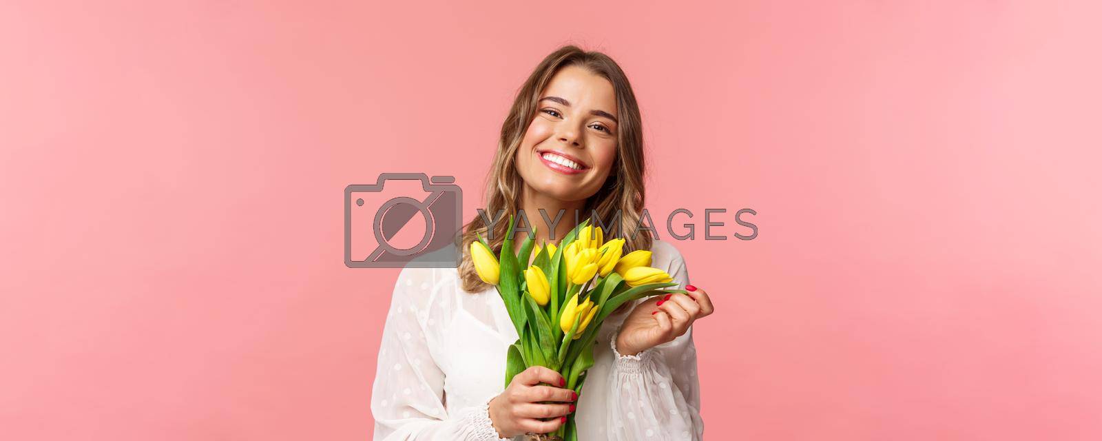 Spring, happiness and celebration concept. Close-up of lovely happy young blond girl in white dress, daydreaming after perfect date, holding yellow tulips and smiling at camera, pink background.