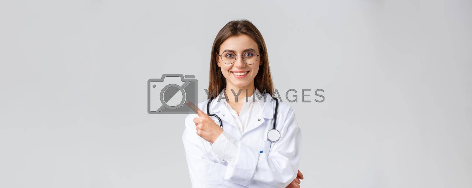 Healthcare workers, medicine and covid-19 pandemic concept. Good-looking professional female doctor in white scrubs and glasses, pointing fingers left, smiling friendly, showing information.