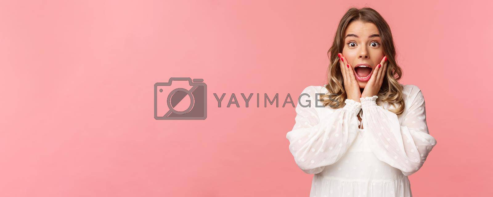 Royalty free image of Happy and surprised blond girl hear awesome news, gasping speechless staring camera with hands on cheeks, being impressed and stunned over great event, standing pink background by Benzoix