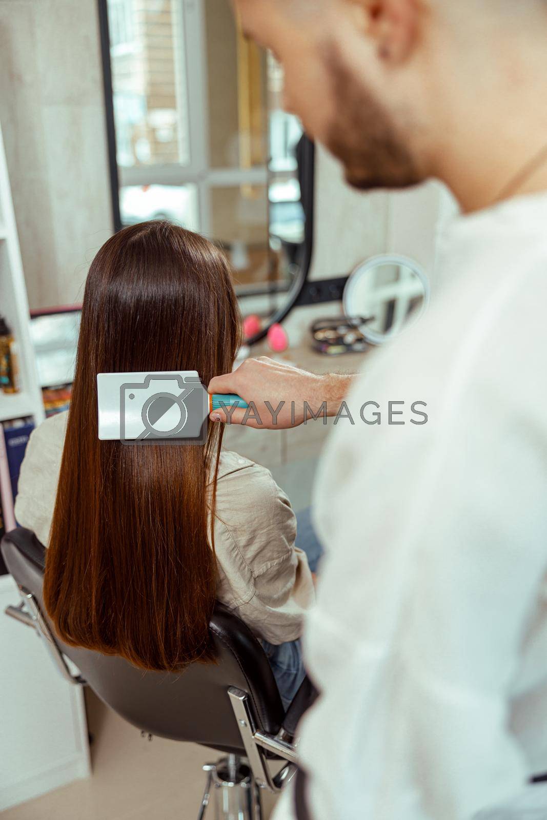 Hairstylist brushing long and sleek brown hair of female client at beauty salon. Hair care