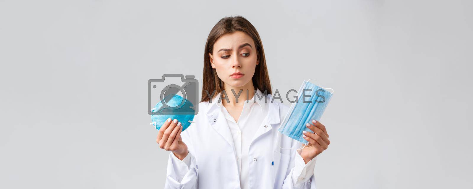 Covid-19, preventing virus, healthcare workers and quarantine concept. Cute female doctor in white scrubs, choosing between medical mask and respirator, pick personal protective equipment.