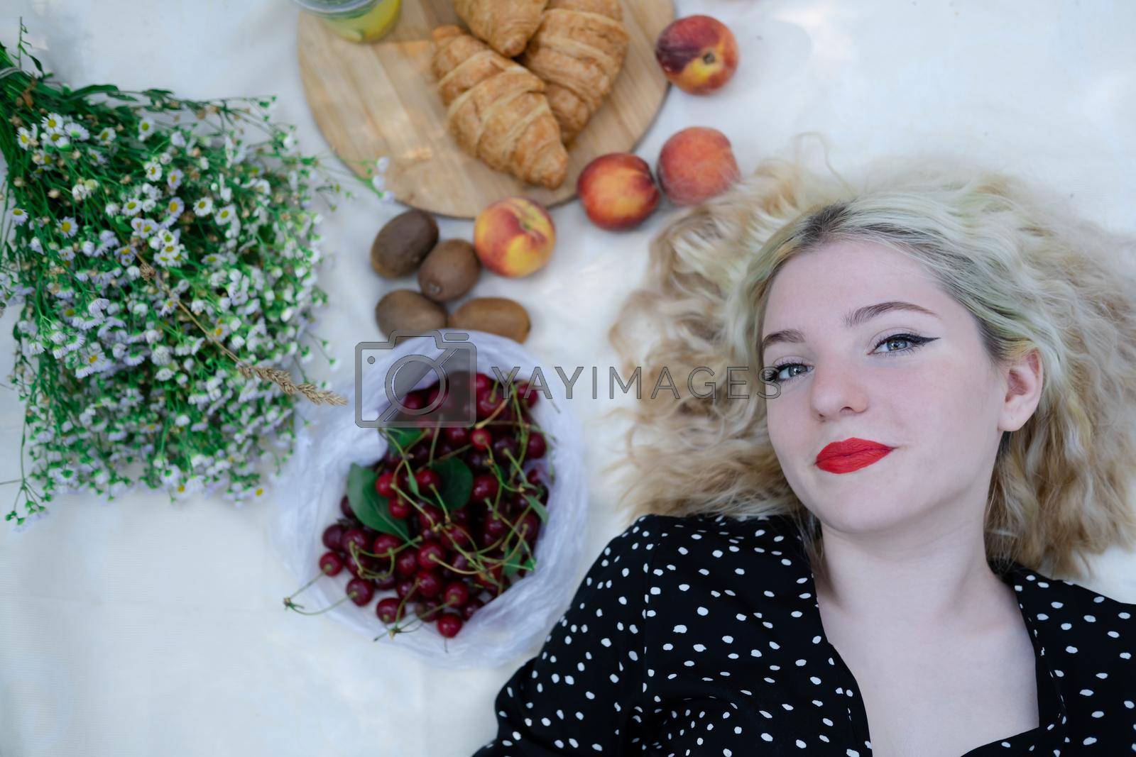 sweet blonde girl on picnic lying on white blanket in park. fresh summer refreshing drink and snacks. fruits and flowers.