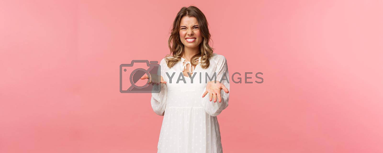 Royalty free image of Portrait of dissatisfied, disappointed cute girl in white dress, cringe show tongue, smell something awful, pointing with raised hands at bad unappropriate thing, grimacing dislike, pink background by Benzoix