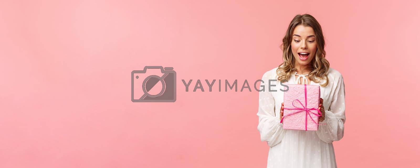 Holidays, celebration and women concept. Portrait of surprised charming young blond girl receive surprise gift, holding present in pink box look at it amused, curious whats inside, studio background.