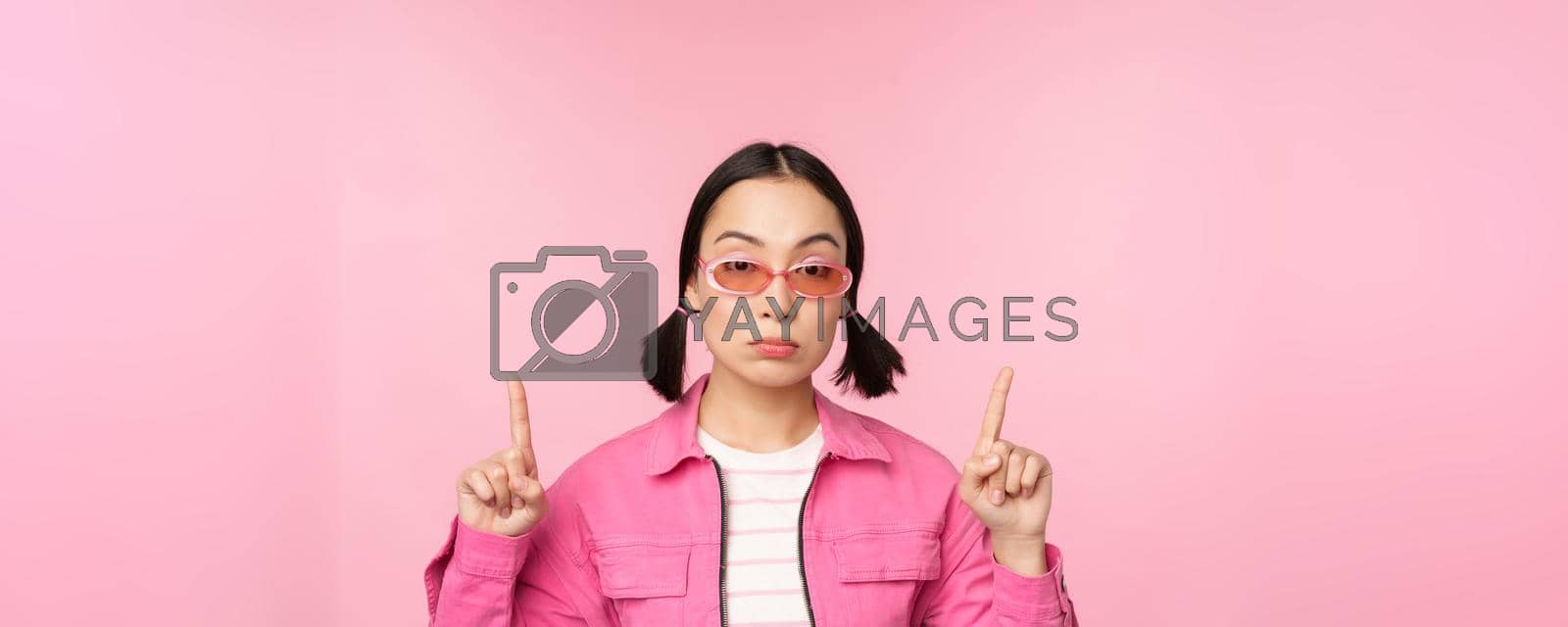 Royalty free image of Close up portrait of asian stylish girl, looking skeptical, pointing finger up with unamused face, standing over pink background by Benzoix