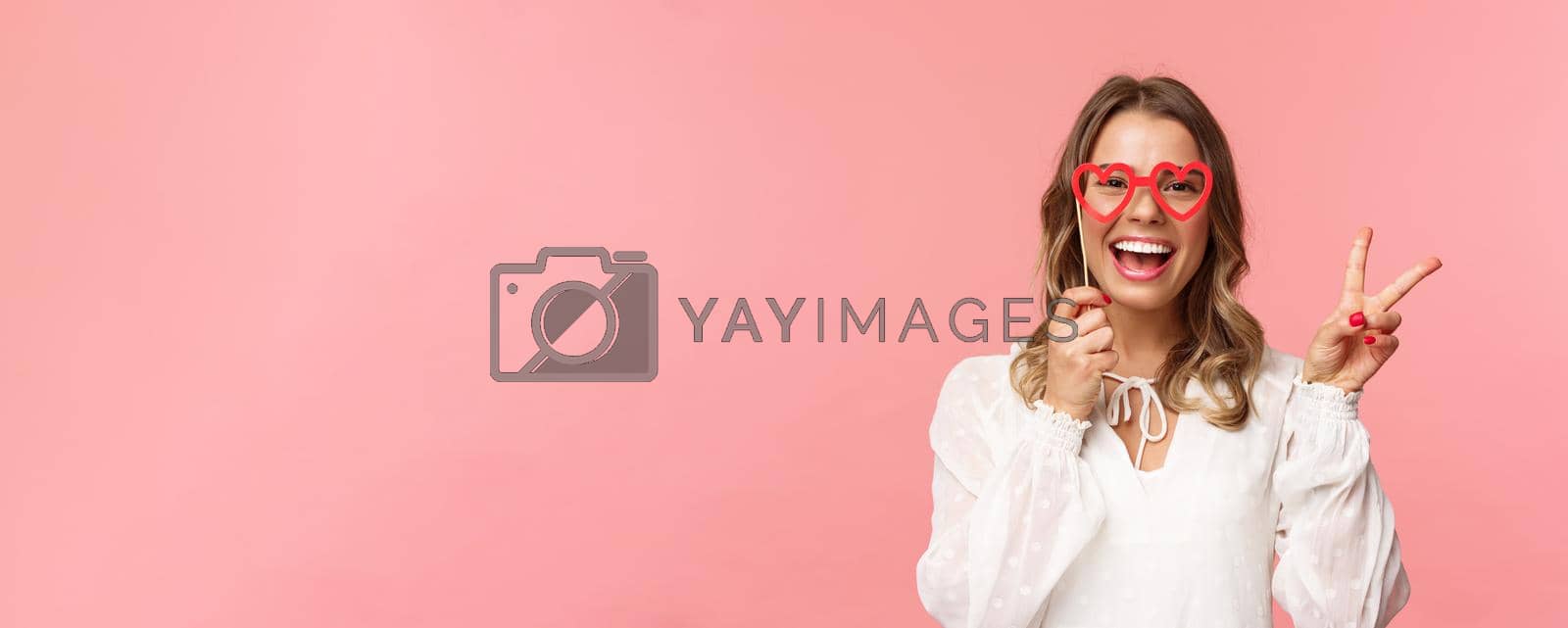 Spring, happiness and celebration concept. Close-up portrait of upbeat blond girl at party, wear white dress holding heart-shaped glasses mask over eyes and make peace sign cheerful.