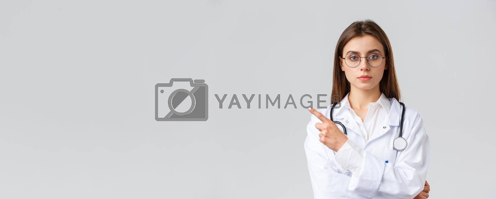 Healthcare workers, medicine, insurance and covid-19 pandemic concept. Serious-looking professional female doctor, nurse in white scrubs and glasses pointing finger left at banner or clinic info.