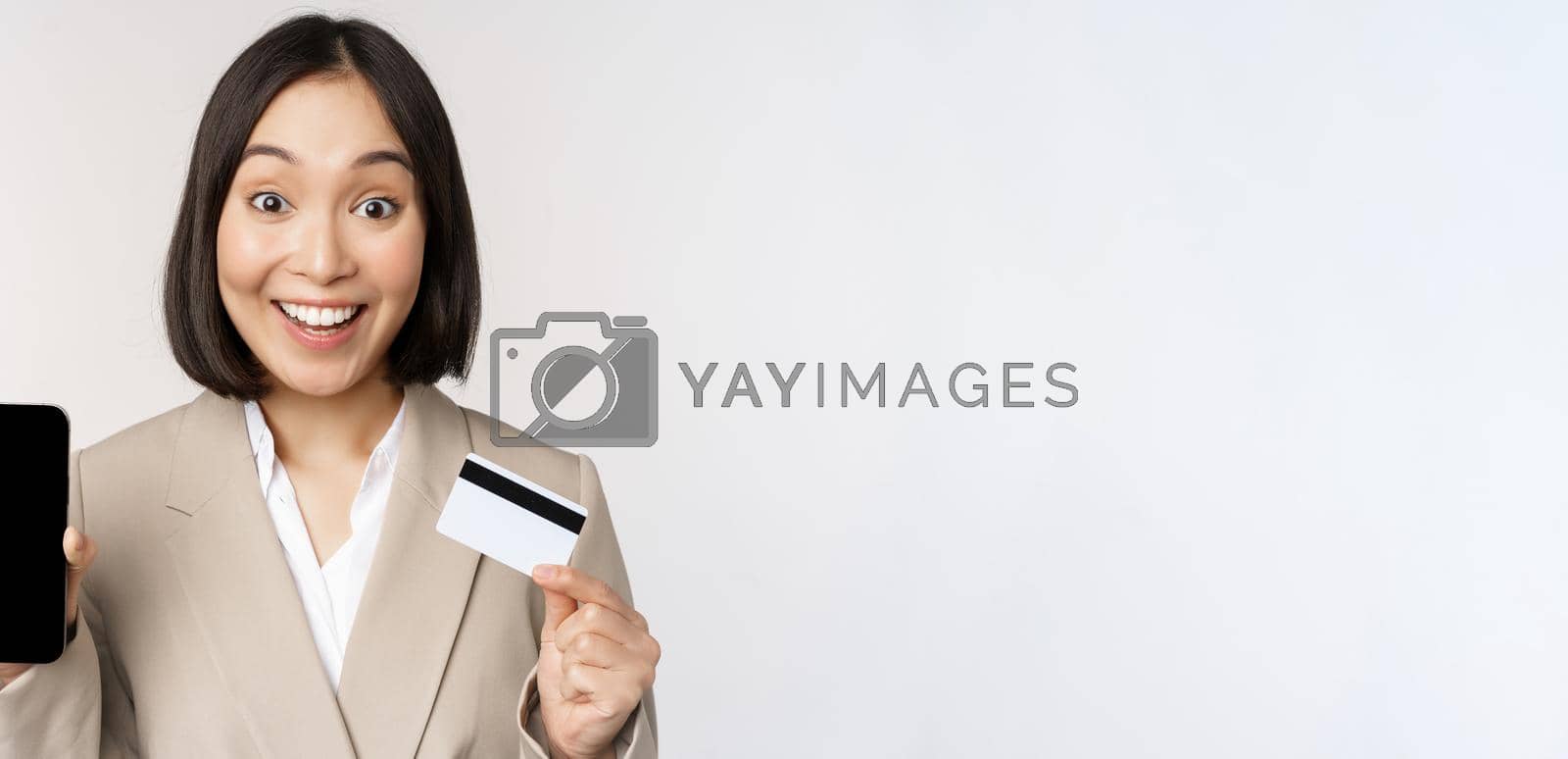 Royalty free image of Corporate woman with happy, enthusiastic face, showing credit card and smartphone app screen, standing in suit over white background by Benzoix