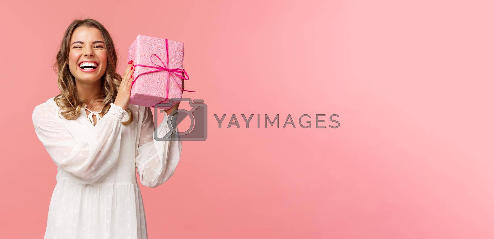 Holidays, celebration and women concept. Portrait of happy charismatic blond girl shaking gift box wondering whats inside as celebrating birthday, receive b-day presents, pink background.