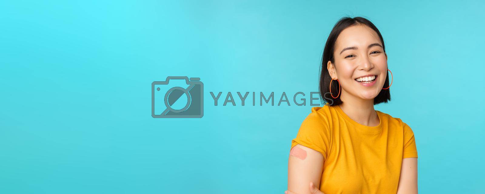 Royalty free image of Vaccine campaign from covid-19. Happy and healthy asian girl laughing after vaccination from coronavirus, bandaid on shoulder, wearing yellow t-shirt, blue background by Benzoix