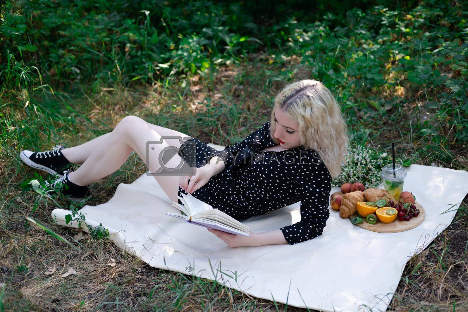 Royalty free image of portrait of young woman on a picnic on plaid in park reading a book with tasty snacks by oliavesna