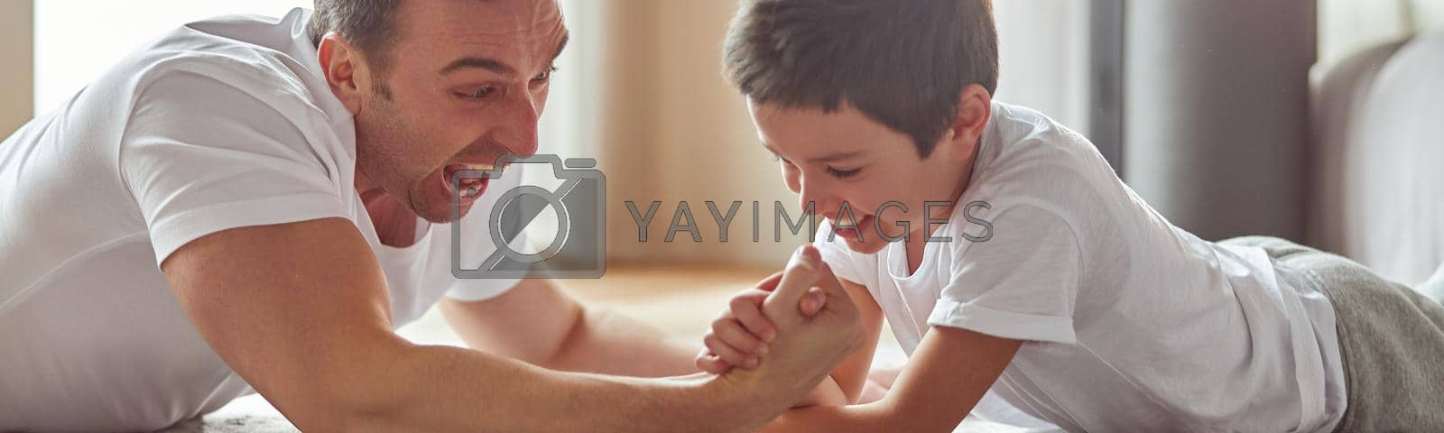 Jolly father is having fun with little boy on floor in living room and doing armwrestling battle