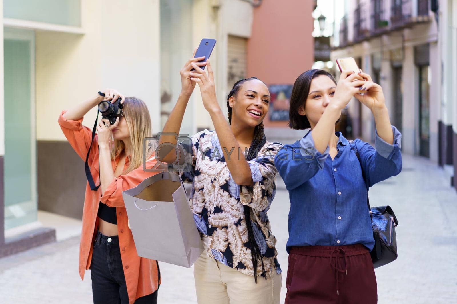 Group of glad diverse female friends taking pictures of street with smartphones and photo camera while standing on sidewalk near buildings