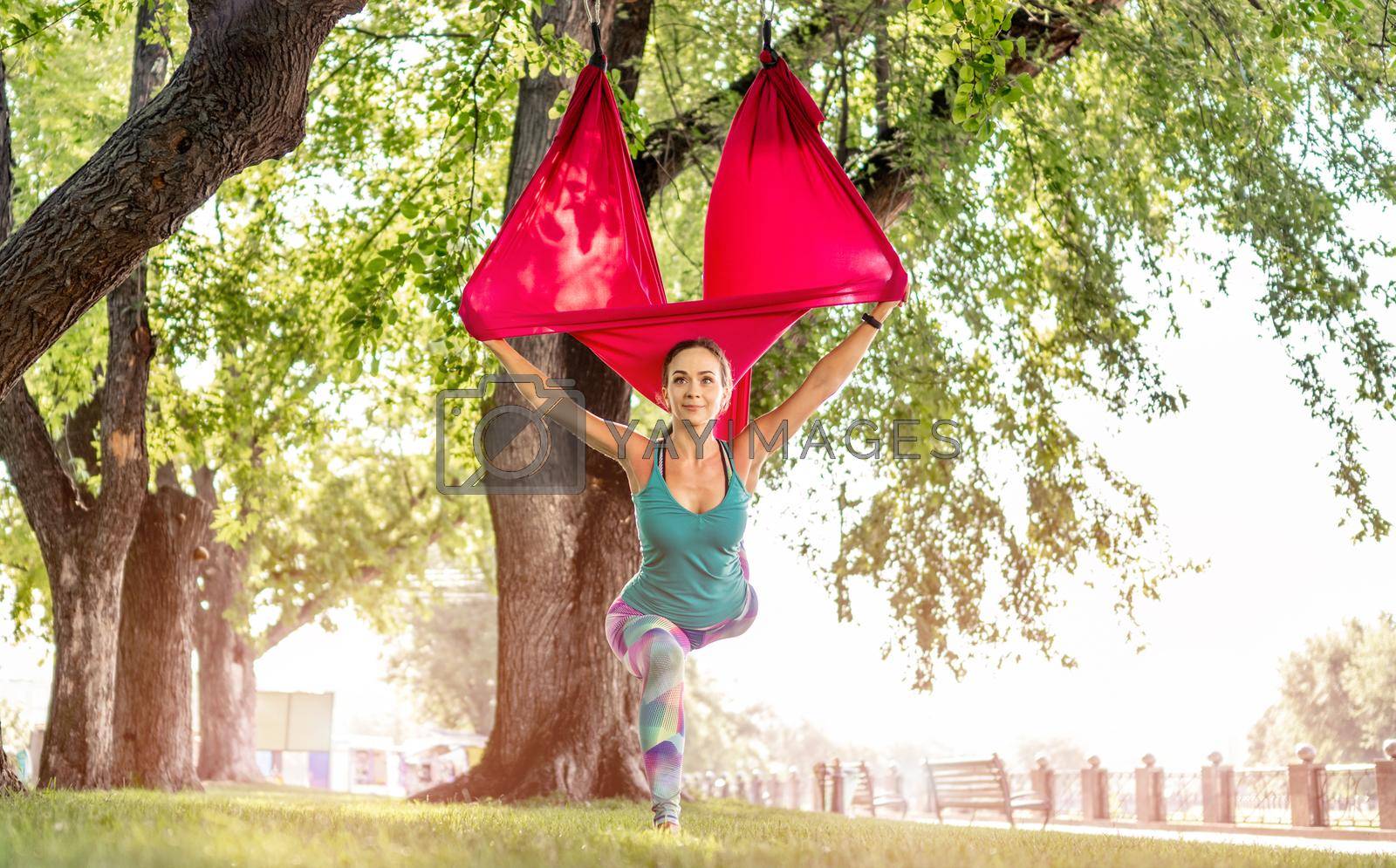 Sport girl practicing fly yoga in hammock at nature and stretching her body standing on one leg. Active woman during aero gymnastics outdoors in summer