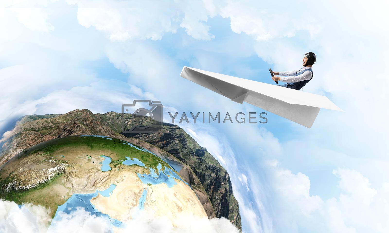 Pilot in leather helmet and goggles driving paper plane on background of blue cloudy sky. Traveling around the world. Funny man flying in small paper airplane. Earth horizon with high mountain range