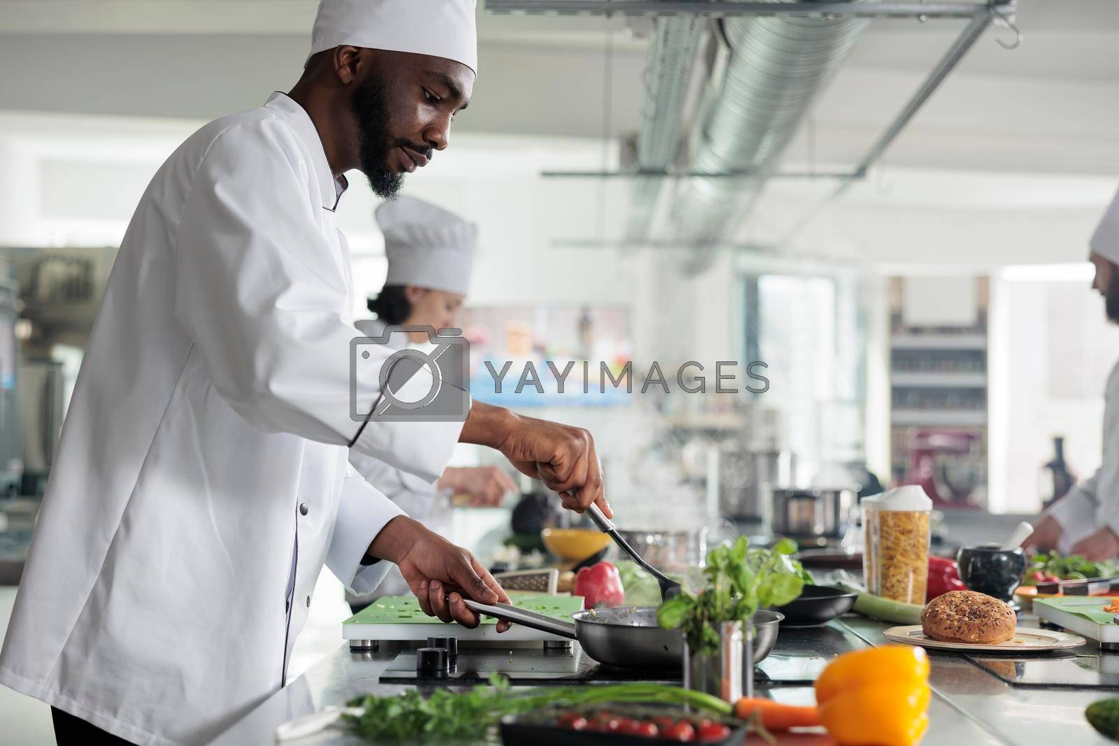 Food industry worker preparing meal in restaurant professional kitchen. Skilled head chef stirring with spatula in pan while cooking gourmet dish for dinner service, garnished with fresh herbs.