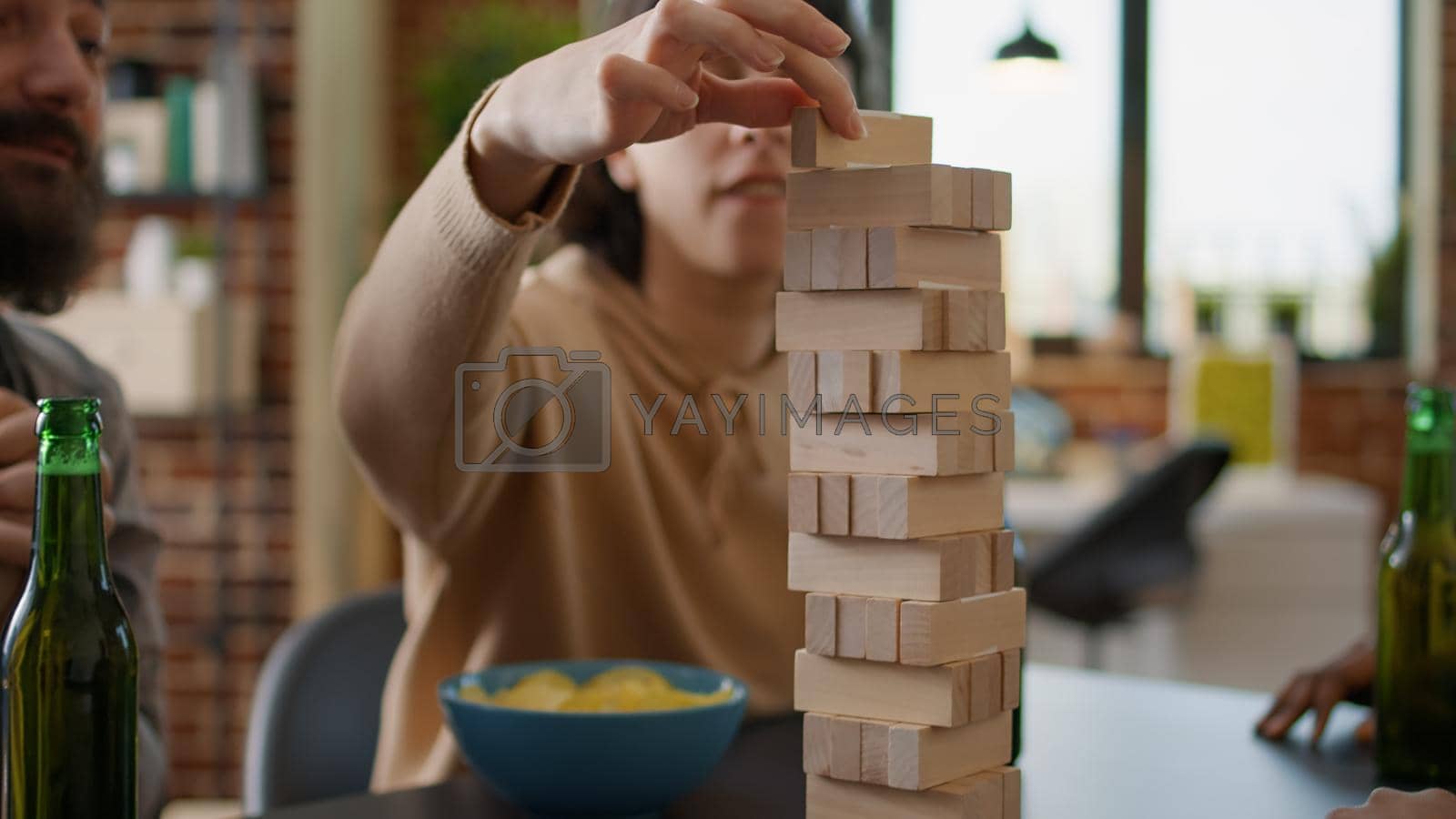 Happy people having fun with wooden tower on table, playing with building blocks structure and square pieces together. Friends enjoying society games pasttime activity laughing.