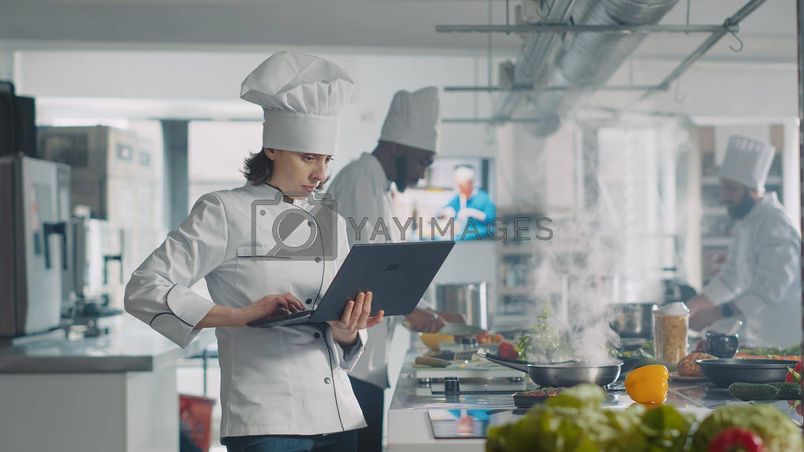 Female cook looking at laptop screen to follow food recipe, making professional gourmet dish. Woman in uniform cooking gastronomy meal with organic ingredients, using computer in kitchen.