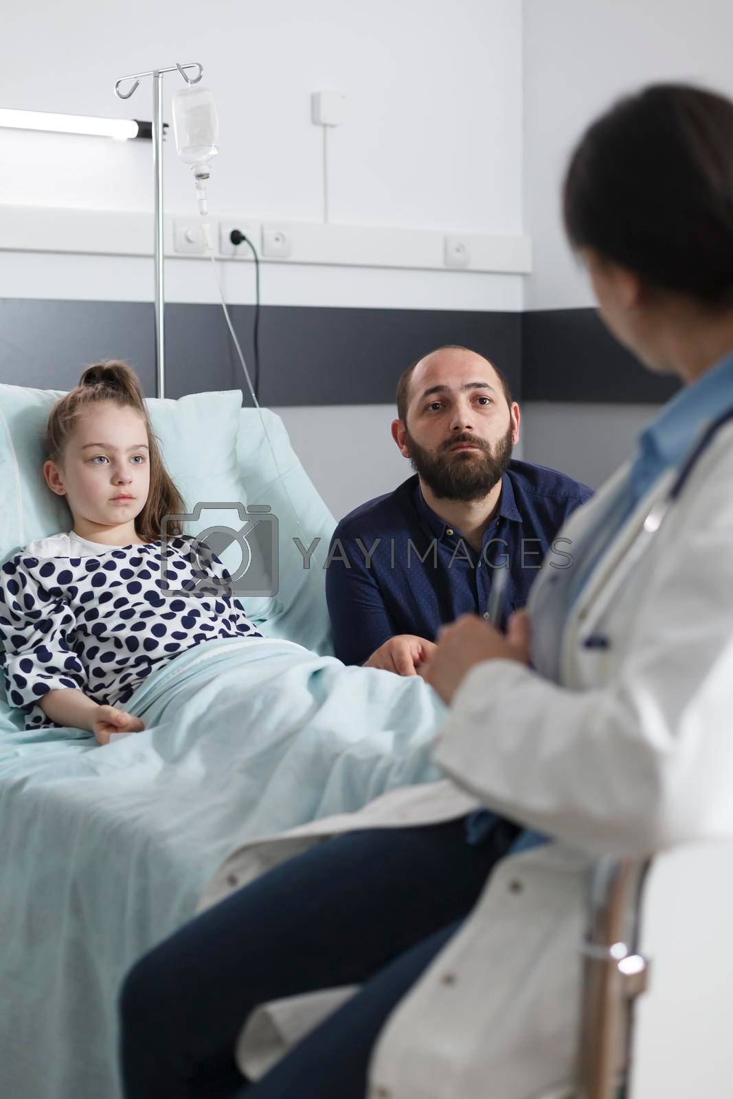 Healthcare hospital pediatrician specialist explaining to worried father daughter illness symptoms and viable treatment. Sick little girl resting in pediatric hospital bed in recovery ward room.