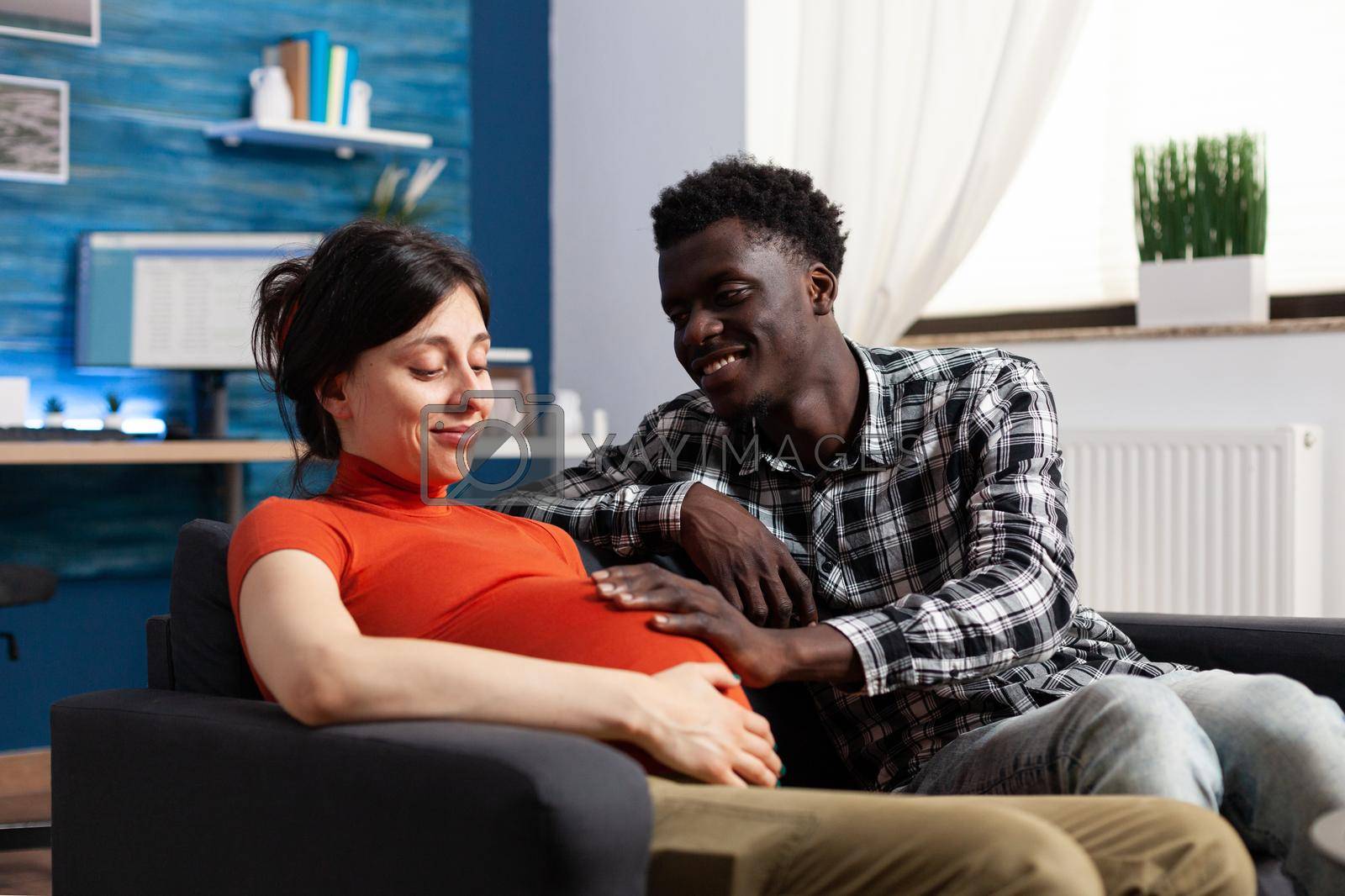 Joyful future father touching pregnant cheerful wife tummy while expecting baby at home. In love interracial young couple waiting for newborn being happy for pregnancy and future parenthood.