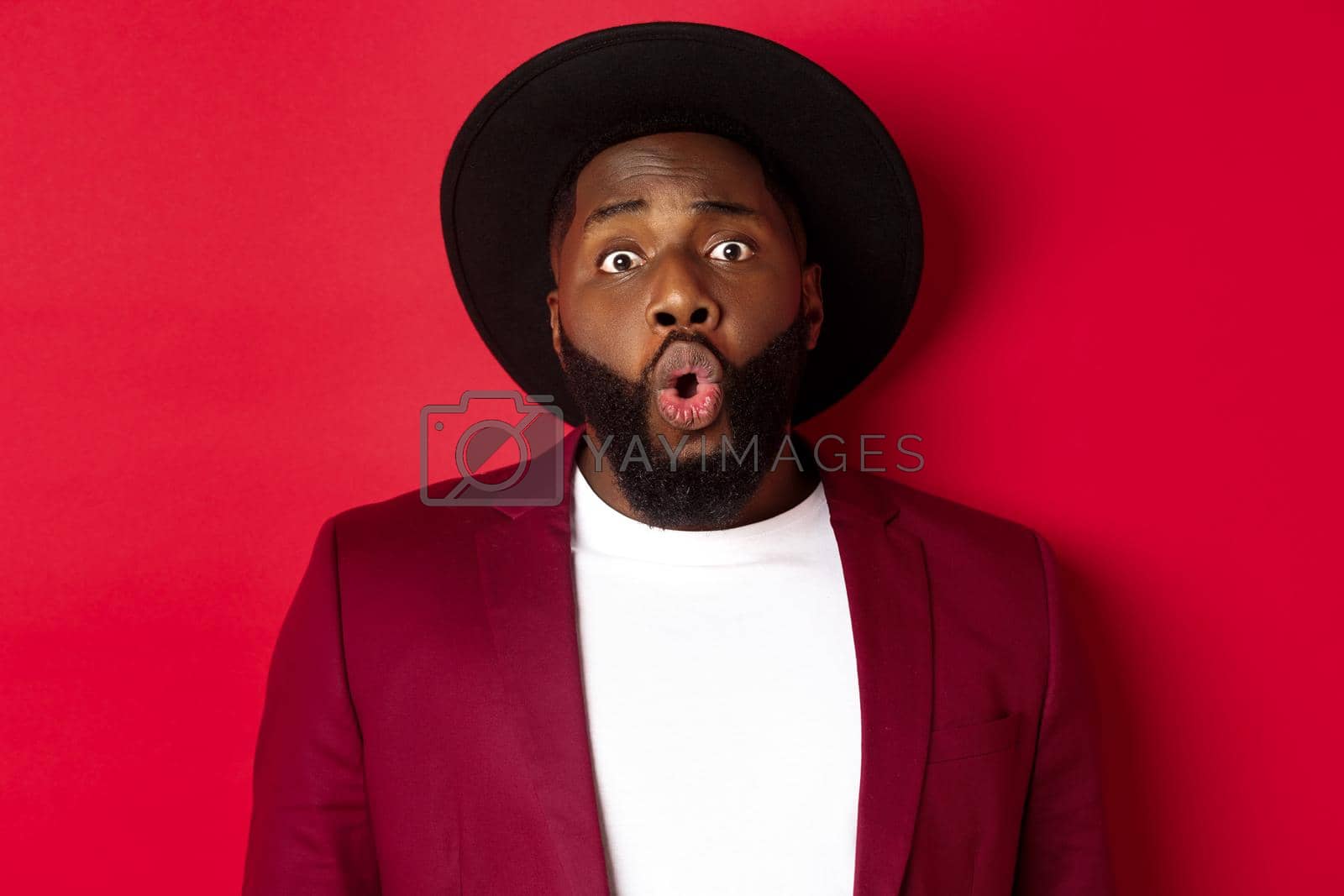 Royalty free image of Close-up of surprised Black man staring at camera with wonder, standing against red background by Benzoix