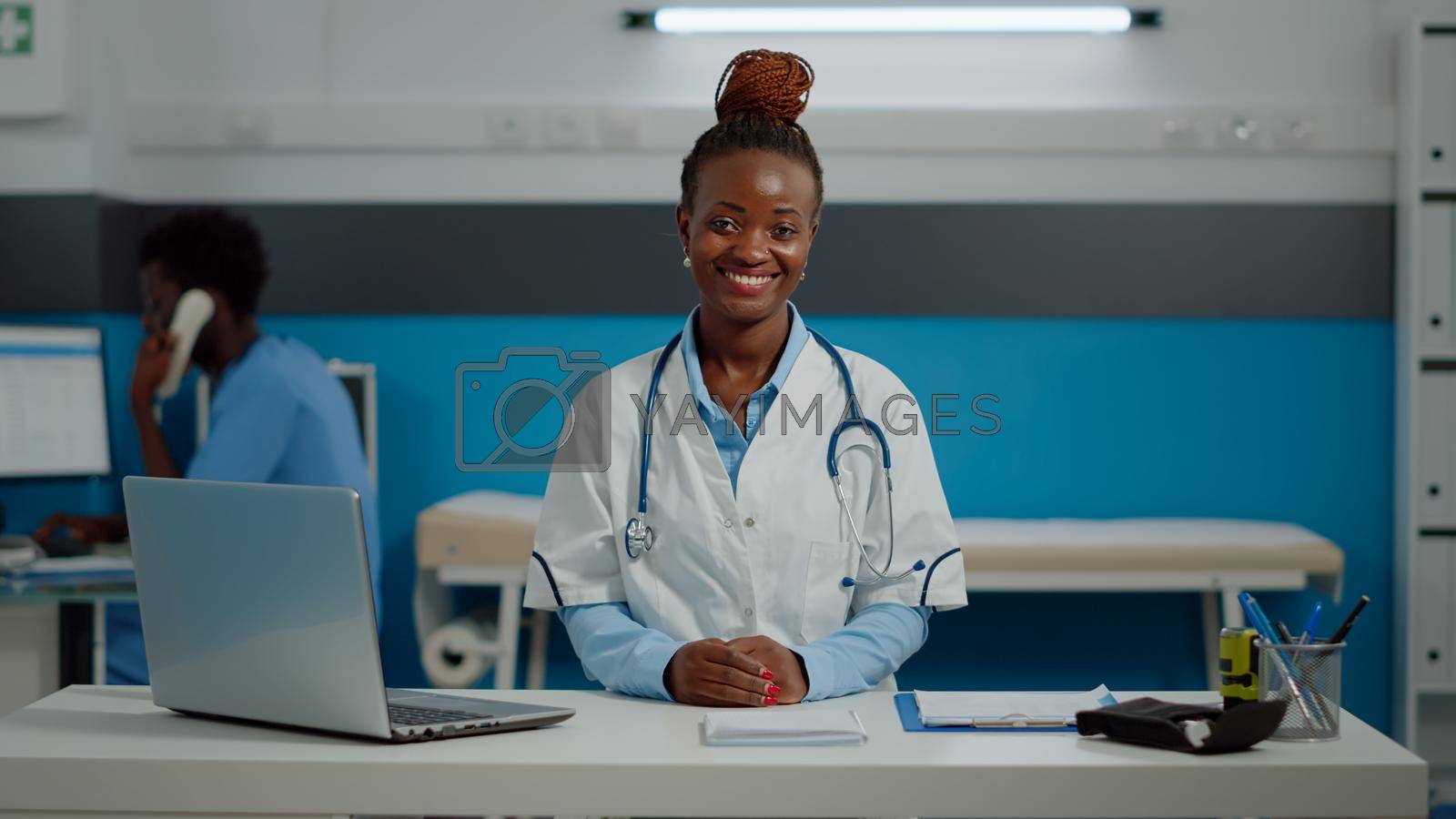 Portrait of woman working as medic in office at healthcare clinic. Doctor wearing white coat and stethoscope while sitting at desk in medical cabinet smiling and looking at camera