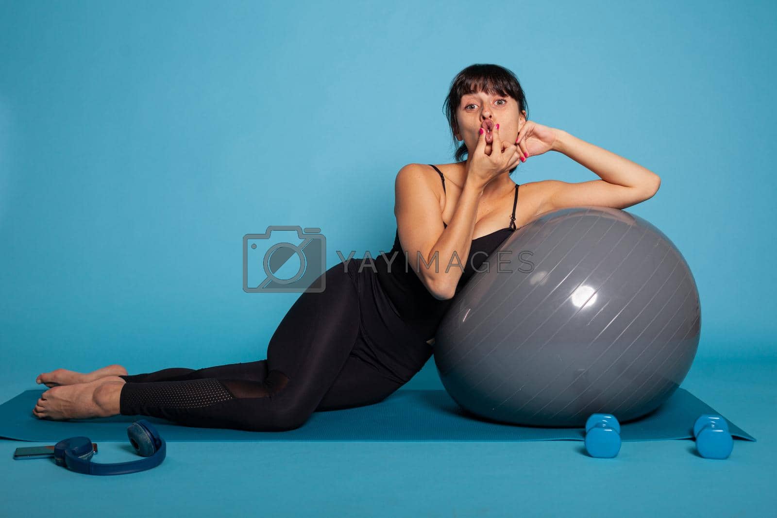 Personal trainer sitting on yoga mat leaning on fitness fitball making funny expression during fitness workout. Active fit woman stretching body muscles working at healthy lifestyle in studio